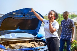 image of beautiful african lady with smart phone in front of an opened bonnet of a car, with guy bit blurred at the back- transportation concept