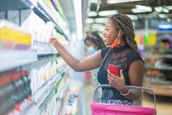 selective focus of cheerful african lady, carrying basket in her arm- black woman in front of shelf, face mask lowered at her chin- shopping concept