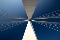 Minimalist abstract architecture shot featuring a white concrete pillar of a suspension bridge with two bunches of suspension cables against a clear blue sky