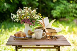 Bouquet of meadow flowers, croissant, cup of tea or coffee, books on table in summer idyllic garden. Rest in garden, reading books, breakfast, vacations in nature concept. Summertime in garden