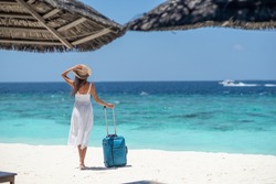 Happy girl in white dress and hat holding suitcase on the beach