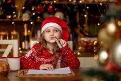 a little cute girl child in a red sweater and a hat in a dark kitchen with a Christmas tree writes a letter to Santa Claus and thinks what to wish for her and waits for the new year or Christmas