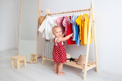 Baby girl stands near a wardrobe and chooses a dress and smiles. Dressing closet with clothes arranged on hangers. Wardrobe of newborn, kids, toddlers, babies full of all clothes. montessori wardrobe.