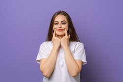 I am not happy with this idea. Studio portrait of gloomy annoyed girl stretching mouth in fake smile with index fingers, standing in blank white t shirt over purple background