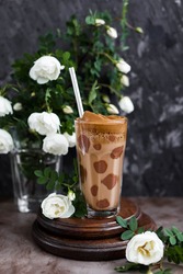 Coffee with chocolate and whipped foam in a transparent glass with a straw on a table with white roses. Unusual design of a coffee drink.