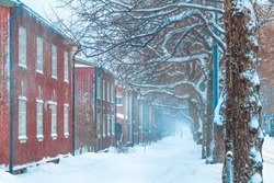 Winter street scene. Old wooden building facades at a street covered by snow. Snowfall in city.  Red houses and trees lined up along snowy footpath. Snowstorm in Helsinki, Finland. Blizzard in town.