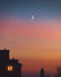 Young crescent moon on the sky over city. Silhouette of towers and houses with chimneys on roofs. Night city buildings with one lonely light in the window with flowerpot. Cityscape. Evening home scene