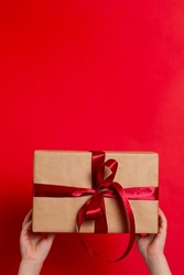 Merry Christmas. Children's hands hold a gift box tied with red ribbon on a red background. Vertical. A place for text. High quality photo
