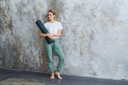 A beautiful blonde girl stands with a twisted rug against an abstract wall. A yoga mat in the hands of a trainer. Sports on the floor. High quality photo