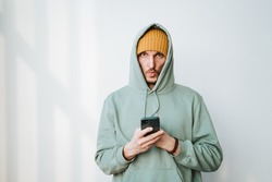 Hipster put a hood on his head, a mobile phone in his hands. Green sweatshirt, yellow hat. Sun glare on the wall. High quality photo