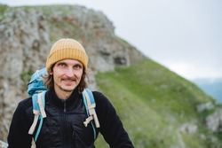 Portrait of a guy in the mountains, a man smiling looks at the camera, a man in a yellow hat in nature, a tourist with a backpack in the mountains, a mountain hike, a smile on his face.
