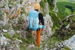 A guy stands with a backpack on a rock looking down, a big blue backpack for things, a hiker on a hike in the mountains view from behind, trekking in the mountains, traveling alone