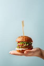 Hand holding burger. Hamburger with meat free plant based cutlet, tomatoes, onions, white sauce and microgreens on blue background. Healthy vegan or vegetarian food concept. Copy space.