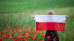 Blond girl holding flag of Poland in the poppy field. Back view. Polish Flag Day. Independence Day. Travel and learn polish language concept. Selective focus.