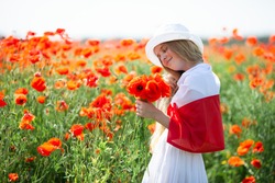 Blond girl covered with flag of Poland holding bouquet of poppies in the poppy field. Polish Flag Day. Independence Day. Love Poland concept. Selective soft focus.
