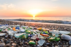 Colored sea glass with beach pebbles and shells in the mediterranean coast and in the background sea and waves with sunset and sky, sandy beach with coast glass, beach glass.