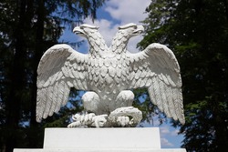 White doubleheaded metal eagle, looking in different directions, against the background of trees and blue sky with clouds.