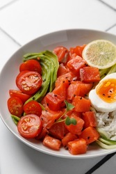 Poke bowl with salmon, avocado, cherry, egg and rice on white tile background with chopsticks. Top view