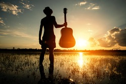 Asia hipster man.Musician holding acoustic guitar and standing in water at sunset