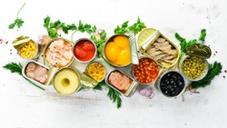 Food background in tin cans. Canned vegetables, beans, fish and fruits on a white wooden background. Top view.