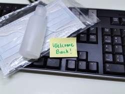 Welcome back note with hand sanitizer and mask on work keyboard; Back to work note with alcohol gel to prevent coronavirus / infection