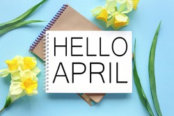 Hello April . text on white notepad paper on blue background. near notepad with yellow flowers and green leaves