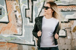Girl wearing t-shirt, glasses and leather jacket posing against street , urban clothing style. Street photography