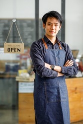 Portrait of young small business owner standing with crossed arm in coffee shop. Happy handsome asian owner man in casual wearing denim apron standing in coffee.Looking at the camera.