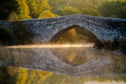 Roman stone bridge, with reflection in the water and sun rays entering from below and with fog, mist