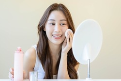 Happy beauty, beautiful asian young woman, girl looking in to mirror, holding cotton pad, applying facial wipe on her face, removing makeup before shower in bathroom, skin care on white background.