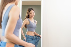 Shape slender, thin waist, attractive slim asian young woman, beautiful girl hand show shape her weight loss, wearing in big, large or over size jeans and looking into mirror. People body fit healthy.