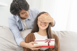 Celebrating anniversary, surprise. Relationship asian young couple love, boyfriend use hand cover girlfriend eye, give a gift by hide box at behind, woman getting present while sitting on sofa at home