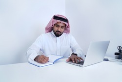 Saudi Arab man writing on page in diary while working on laptop    