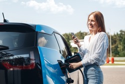 Young beautiful woman traveling by electric car having stop at charging station standing plugging cable browsing internet on smartphone smiling joyful while charing