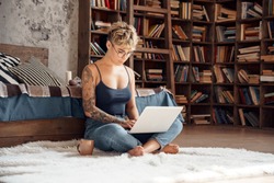 Young woman short hair wearing eyeglasses sitting on fluffy carpet with cup of hot coffee and laptop working online concentrated freelance side hustle