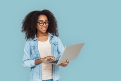 Young african girl wearing eyeglasses standing isolated on gray background holding laptop browsing web looking at screen smiling joyful