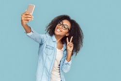 Young african girl wearing eyeglasses standing isolated on gray background taking selfie photo on smartphone posing to camer showing peace gesture smiling happy