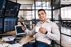 Happy stock broker resting in office, using modern smartphone, drinking beverage and sitting near monitor with cryptocurrency information data on finance market graph