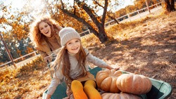 Beautiful young adult mother spending autumn day on farm with her little daughter. Cheerful child choosing small pumpkins, sitting near smiling woman and green wheelbarrow