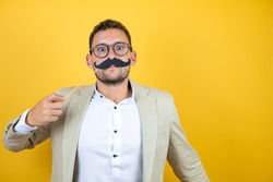 Young handsome businessman wearing suit over isolated yellow background happy with mustache mask