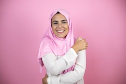Young beautiful arab woman wearing islamic hijab over isolated pink background hugging oneself happy and positive, smiling confident