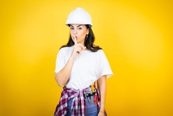 Young caucasian woman wearing hardhat and builder clothes over isolated yellow background asking to be quiet with finger on lips. Silence and secret concept.