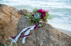 Beautiful colorful wedding bouquet of different flowers with blue and violet ribbon. Pink roses, violet peonies. Ceremony in sea style,decorated at sea shore. Composition on stone, with view at water.