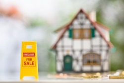Foreclosures and foreclosed home for sale property listings, financial concept : Yellow warning sign board with the words FORECLOSURE FOR SALE, a two-story half-timbered model house, coins on a table.