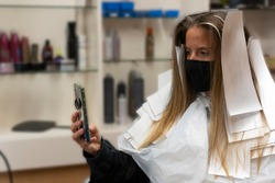 Beautiful woman client in hairdressing salon using smartphone,taking a selfie ,wearing medical mask against coronavirus and hair dye paper for coloring highlights treatment.New normal,Covid19 concept.