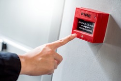 Male hand pointing at red fire alarm switch on concrete wall in office building. Industrial fire warning system equipment for emergency.