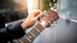 Male hand guitarist adjusting pegs on acoustic guitar during music lesson at home. String musical instrument concept