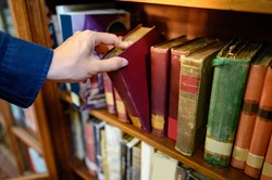 Male university student hand choosing and picking vintage book from old wooden bookshelf in college library. Antique textbook resources for education research. History, Law and Literature learning
