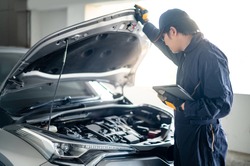 Asian auto mechanic holding digital tablet checking car engine under bonnet hood in auto service garage. Mechanical maintenance engineer working in automotive industry. Automobile servicing and repair
