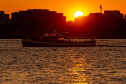 A sunset image featuring sun setting behind buildings, while a river cruise boat with passengers standing on deck is traveling in Potomac river that runs between Washington DC and virginia